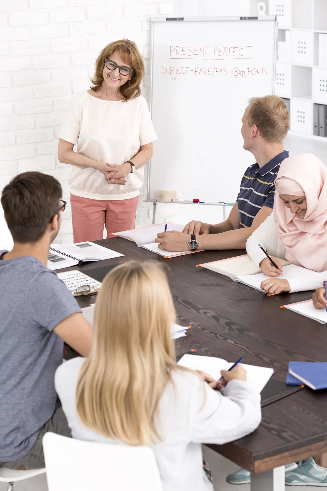 A teacher at a whiteboard with a group of adult learners seated around a table