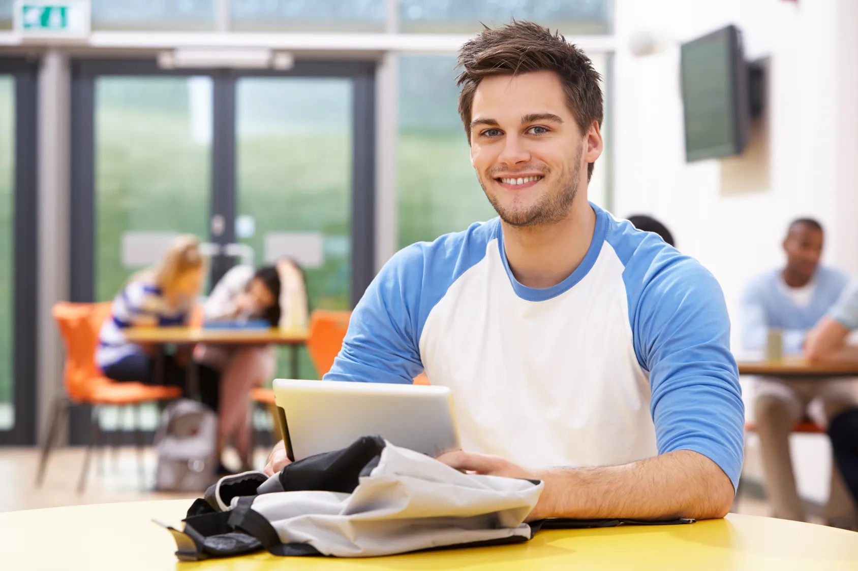 A smiling young man holding a tablet in a school common area