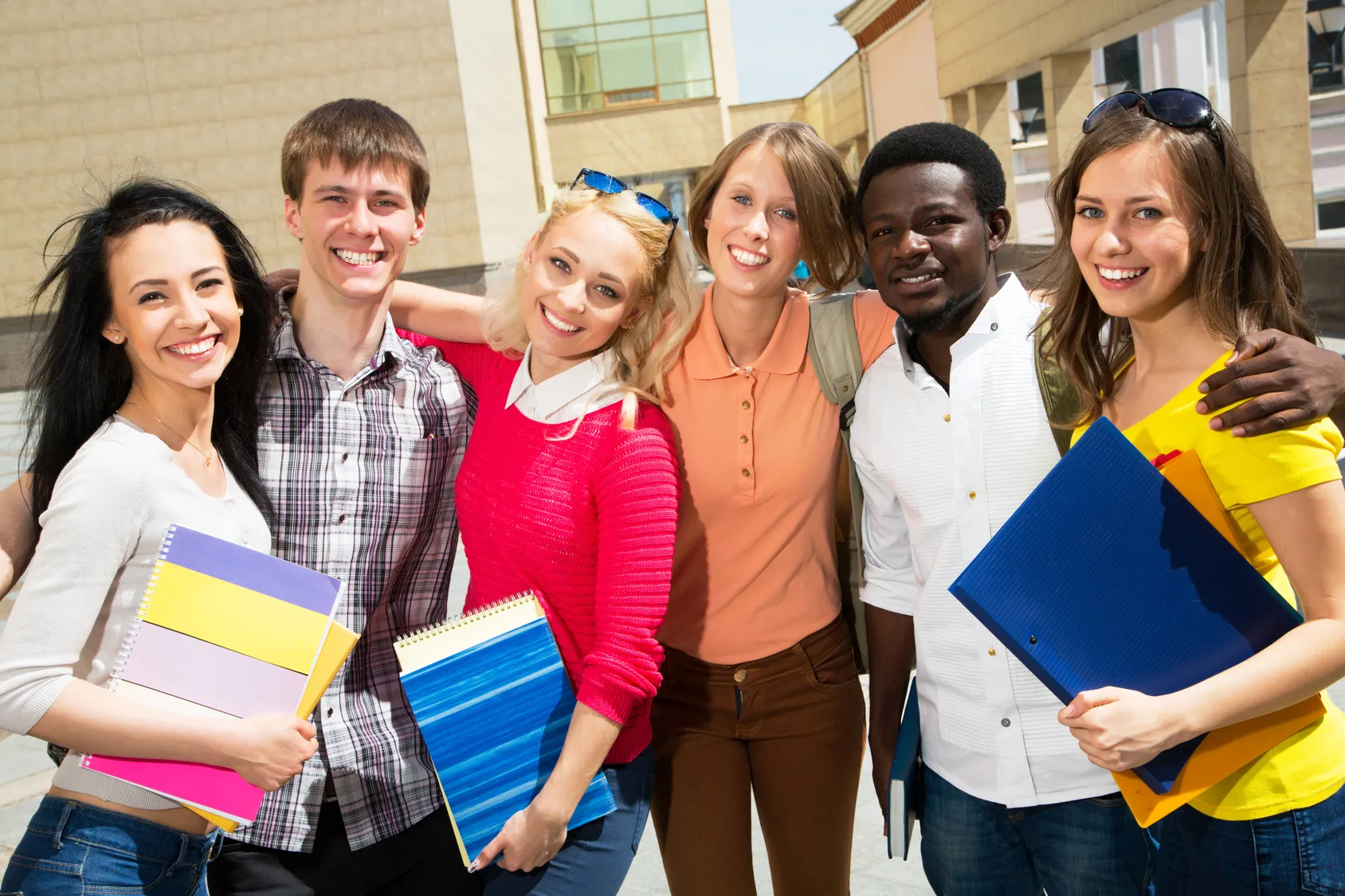Group of six smiling students outdoors in front of a campus building, holding notebooks and folders