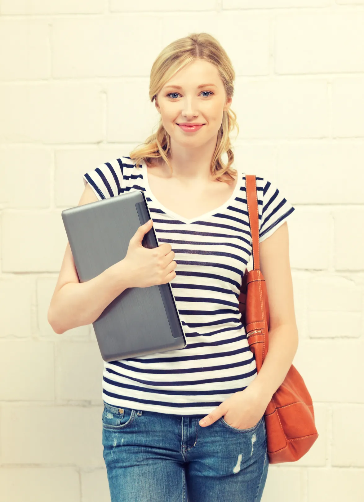 Smiling young woman in a striped top holding a laptop against a white brick wall