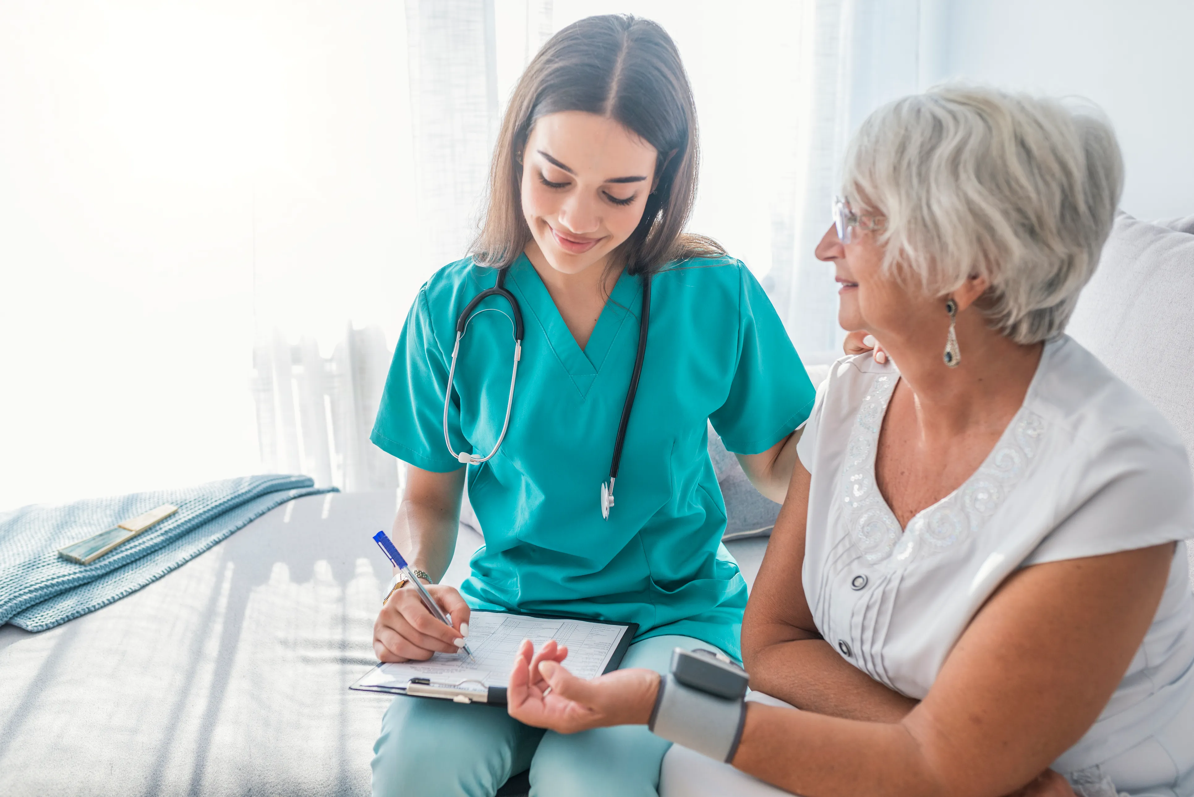 A nurse in scrubs taking notes beside an older patient