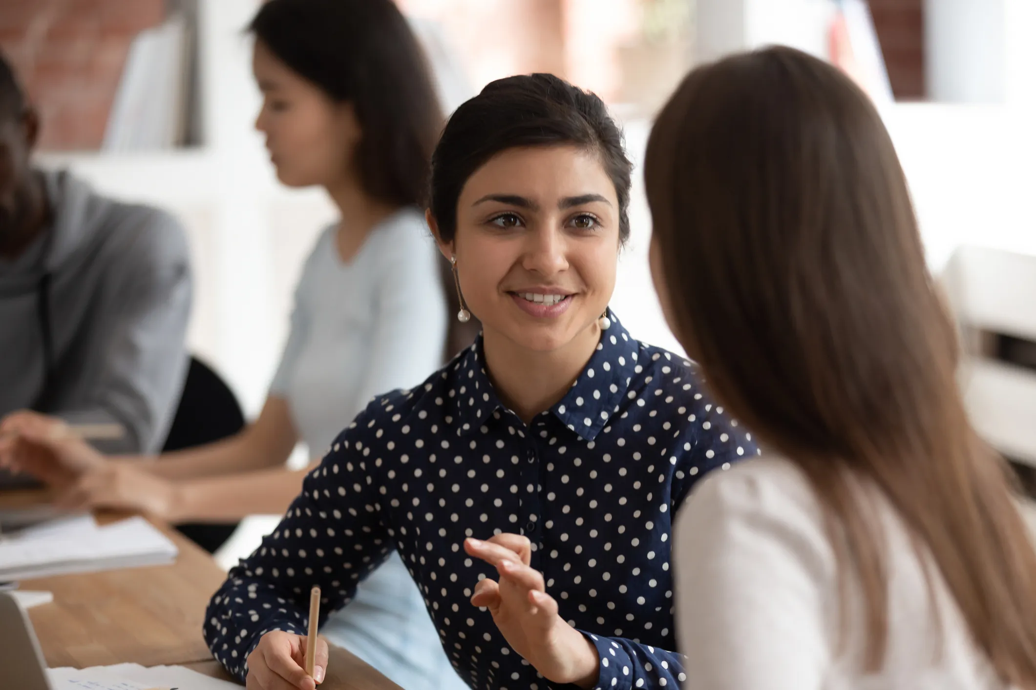 A smiling woman in a polka-dot shirt talking with a colleague at a table