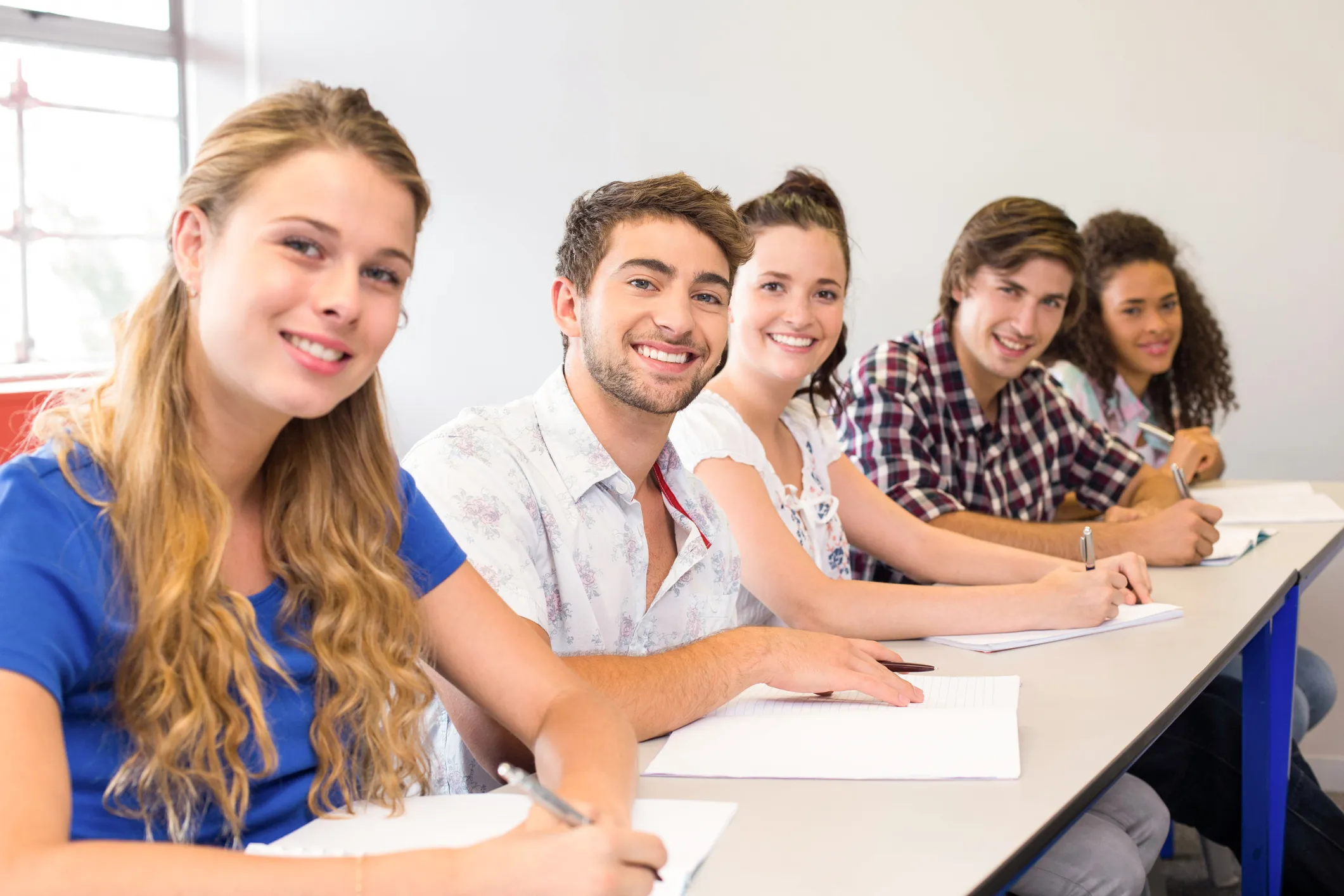 Students seated in a classroom, smiling at the camera beside open notebooks