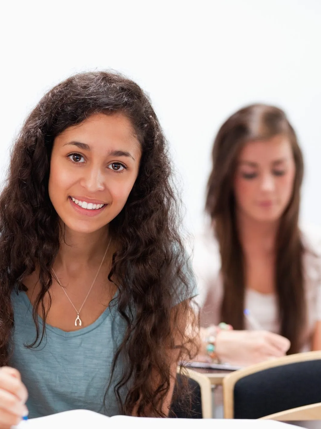 Smiling student with long curly hair at a desk, with a classmate writing notes behind her
